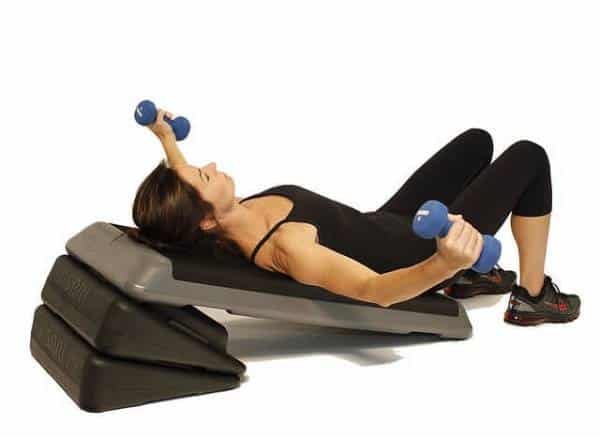 A fit woman works out on an inclined bench, using dumbbells and a stability ball.